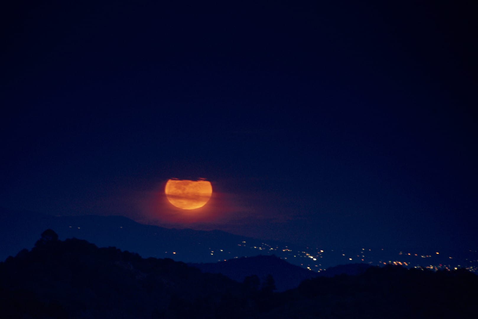 Luna roja elevándose sobre un horizonte montañoso con luces de ciudad al fondo durante la noche.