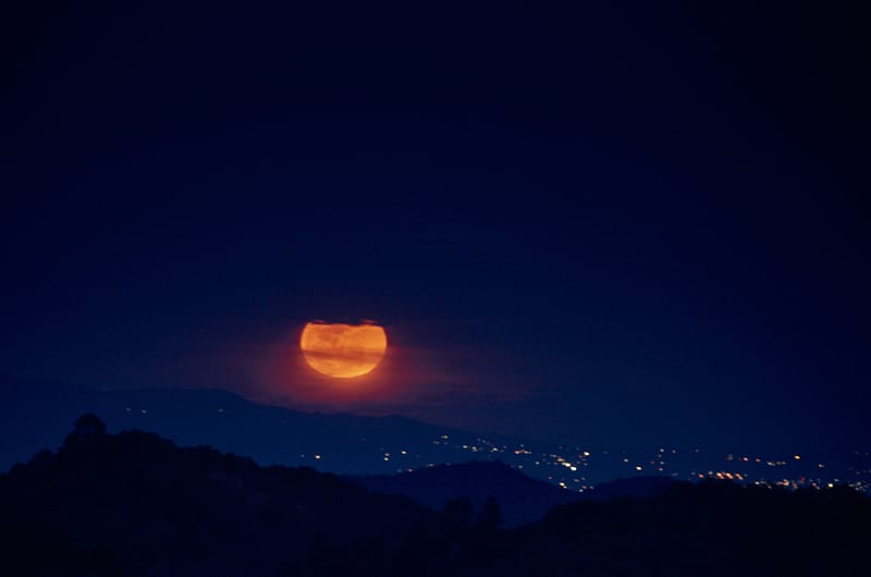 Luna roja elevándose sobre un horizonte montañoso con luces de ciudad al fondo durante la noche.